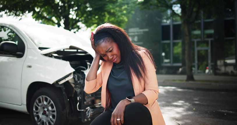 A woman is sitting outside of her crashed vehicle, visibly upset as she holds her head.