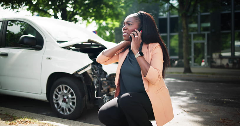 A woman upset on the phone after being in a rideshare car accident. Her damaged vehicle is behind her.