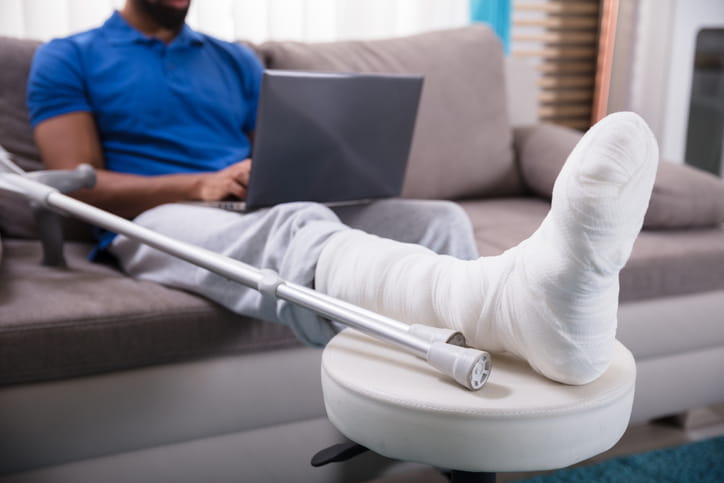 A man with his leg in a cast sits on the couch while he researches what to do after being injured in an Airbnb on his laptop. His crutches are propped up next to him.